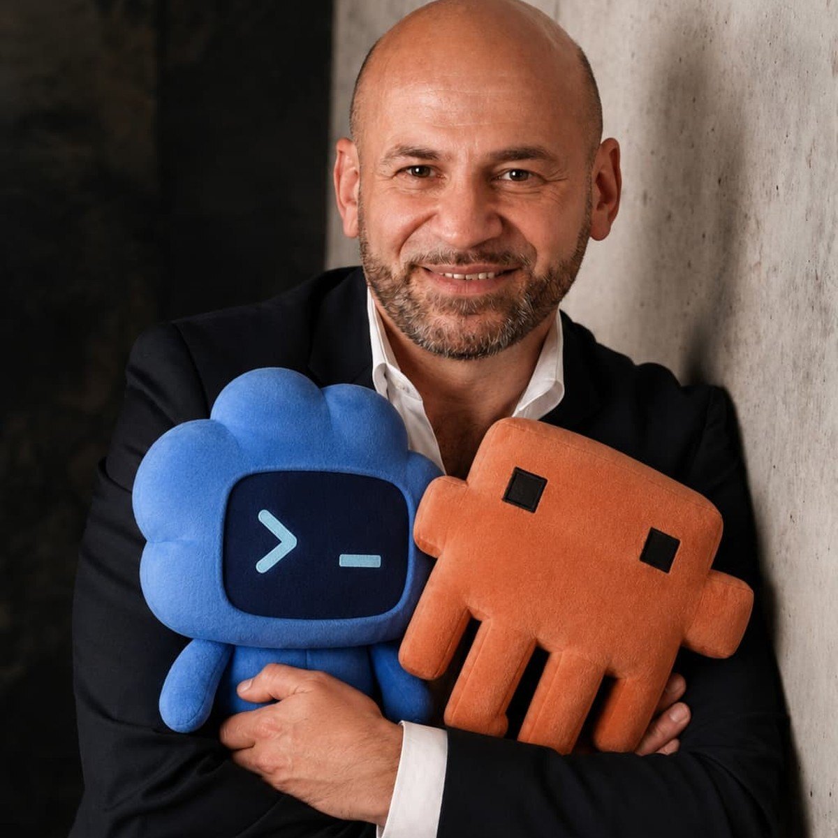 Dr.-Ing. Suat Akyol lehnt an einer Betonwand und hält zwei Plüsch-Maskottchen: links das blaue mit einem Terminal-Prompt für Codex, rechts das orange für Claude.