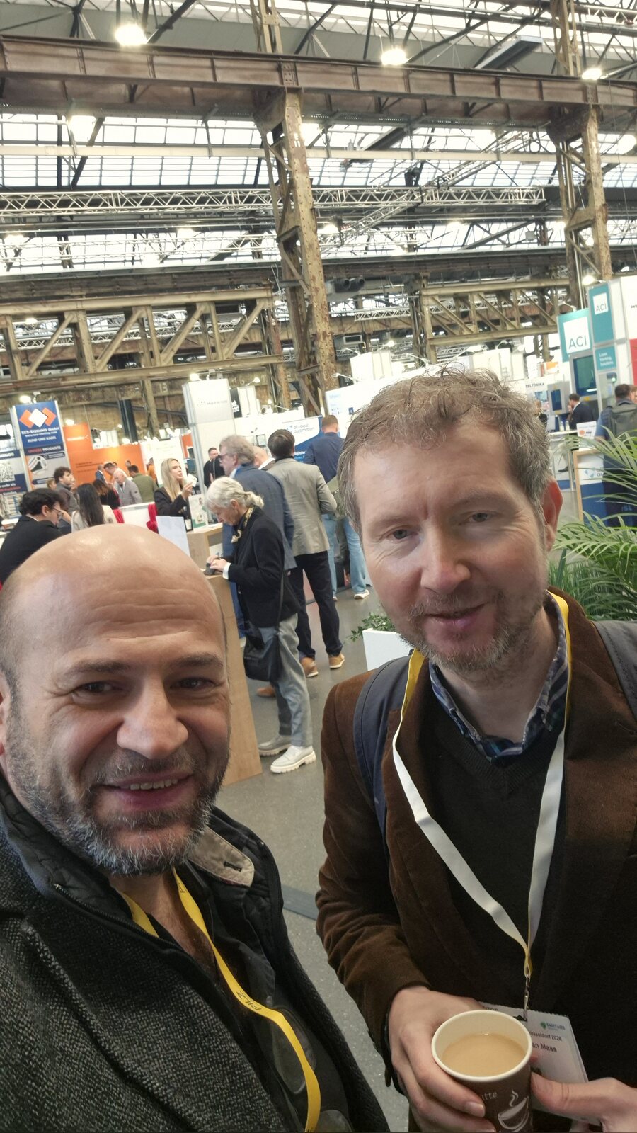 Selfie von Suat Akyol und Stefan Maas mit Kaffeebecher in der Industriehalle des Areal Böhler, im Hintergrund Messestände