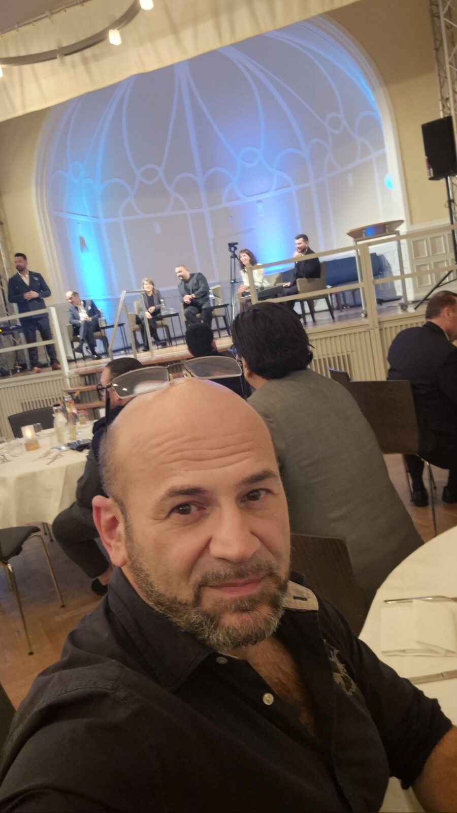 Selfie von Suat Akyol an einem Tisch im Kuppelsaal des Steeler Stadtgartens, im Hintergrund das Podium vor dem blau beleuchteten Rundbogenfenster