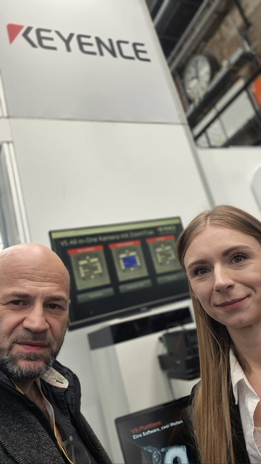 Selfie von Suat Akyol und Sabrina Zurel am Keyence-Stand, im Hintergrund ein Bildschirm mit Prüfbildern der VS-Kamera